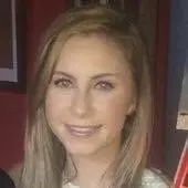A woman with long, straight, light brown hair and light skin smiles at the camera. She is wearing a white top and stands in front of a dark background that includes a colorful wall and a framed picture. Her radiant look is courtesy of the best hair salon Cary NC has to offer.