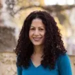 A woman with long, straight, light brown hair and light skin smiles at the camera. She is wearing a white top and stands in front of a dark background that includes a colorful wall and a framed picture. Her radiant look is courtesy of the best hair salon Cary NC has to offer.