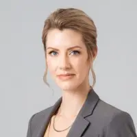 A woman with long, straight, light brown hair and light skin smiles at the camera. She is wearing a white top and stands in front of a dark background that includes a colorful wall and a framed picture. Her radiant look is courtesy of the best hair salon Cary NC has to offer.