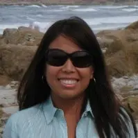 A woman with long, straight, light brown hair and light skin smiles at the camera. She is wearing a white top and stands in front of a dark background that includes a colorful wall and a framed picture. Her radiant look is courtesy of the best hair salon Cary NC has to offer.