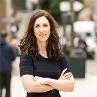 A woman with long, straight, light brown hair and light skin smiles at the camera. She is wearing a white top and stands in front of a dark background that includes a colorful wall and a framed picture. Her radiant look is courtesy of the best hair salon Cary NC has to offer.