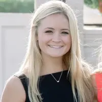 A woman with long, straight, light brown hair and light skin smiles at the camera. She is wearing a white top and stands in front of a dark background that includes a colorful wall and a framed picture. Her radiant look is courtesy of the best hair salon Cary NC has to offer.