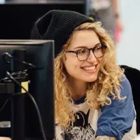 A woman with long, straight, light brown hair and light skin smiles at the camera. She is wearing a white top and stands in front of a dark background that includes a colorful wall and a framed picture. Her radiant look is courtesy of the best hair salon Cary NC has to offer.