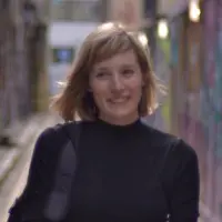 A woman with long, straight, light brown hair and light skin smiles at the camera. She is wearing a white top and stands in front of a dark background that includes a colorful wall and a framed picture. Her radiant look is courtesy of the best hair salon Cary NC has to offer.