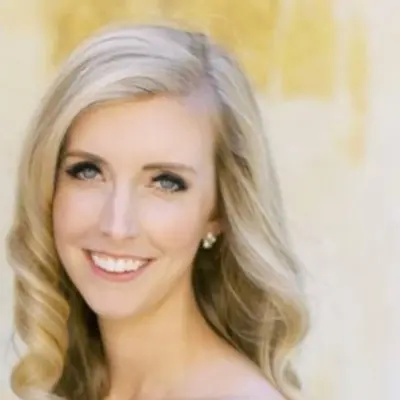 A woman with long, wavy blonde hair and blue eyes is smiling at the camera. She is wearing a pair of small, stud earrings and is posed in front of a light-colored, softly lit background. Her elegant hairstyle was expertly done at a nearby hair salon.