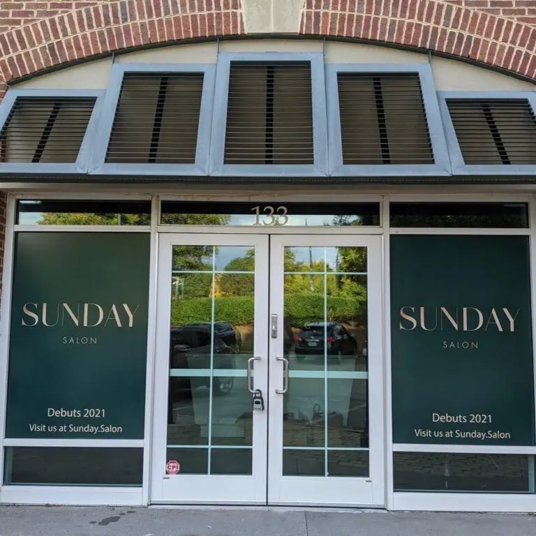 Photo of a storefront with large green windows bearing the text "Sunday Salon" in a modern font. The bottom windows read, "Debuts 2021 Visit us at Sunday.Salon." The building's address, 133, is displayed above the glass doors. The sidewalk is visible in the foreground. best hair salon in Raleigh, NC!