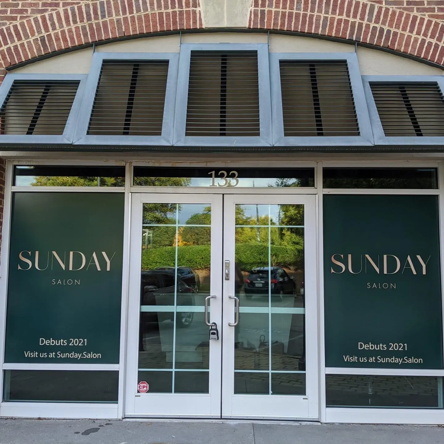 Photo of a storefront with large green windows bearing the text "Sunday Salon" in a modern font. The bottom windows read, "Debuts 2021 Visit us at Sunday.Salon." The building's address, 133, is displayed above the glass doors. The sidewalk is visible in the foreground. Best hair salon Cary NC!