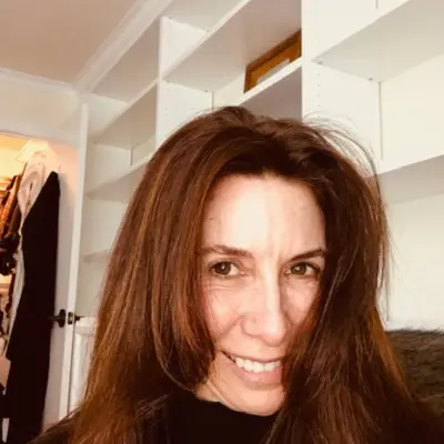 A woman with long brown hair smiles at the camera. She is indoors, standing in front of white shelving units that are partially filled. The background also reveals an open door with clothing visible inside the adjacent room, evoking a cozy vibe akin to a boutique hair salon.