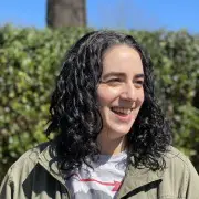 A person with wavy, shoulder-length dark hair, possibly styled by one of the best hair salons in Cary, NC, is smiling while standing outdoors. They are wearing a light-colored jacket over a white shirt. The background features green foliage and a tree, with a clear blue sky above.