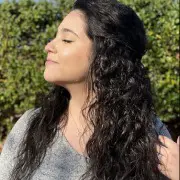 A woman with long, curly dark hair closes her eyes and smiles slightly while facing the sunlight. She stands outdoors against a backdrop of greenery, wearing a grey top, embodying the effortless beauty you'd find at the best hair salon Cary, NC has to offer.
