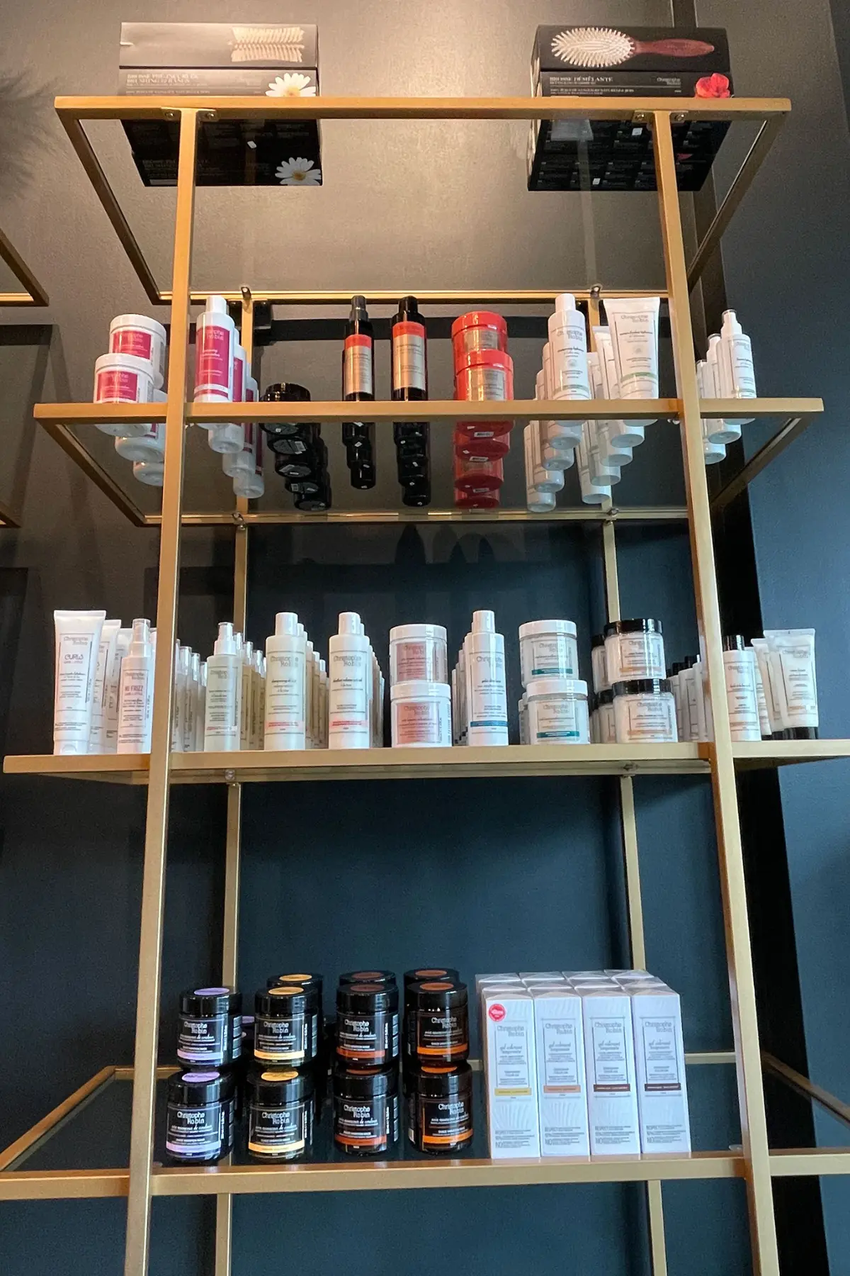 A shelf display with various skincare and beauty products stands elegantly, reminiscent of a Sunday salon. The shelf has multiple levels with items placed in an organized manner, including creams, lotions, serums, and hair brushes. The background is a dark grey wall.