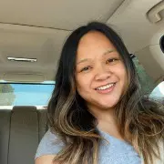 A person with long, wavy hair smiling while seated inside a car. The person is wearing a grey t-shirt, and the car's interior, including the rearview mirror and headrest, is visible. Sunlight is streaming in through the car windows, showcasing hair that looks like it just had a touch-up at Sunday Salon.