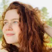 A person with long, curly red hair, styled to perfection by the best hair salon Cary NC, looks off to the side with a serene expression. Their hand is placed behind their head. The background is slightly blurred, showing greenery and trees, suggesting an outdoor setting.