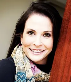 A woman with long dark hair and green eyes is smiling at the camera. She is wearing a colorful scarf and a dark top, standing next to a wooden structure. The background is softly lit, creating a warm and inviting atmosphere, reminiscent of the best hair salon Cary NC has to offer.