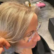 A person with glasses and shoulder-length, grayish-blonde hair is shown from the side. They are indoors in what appears to be the best hair salon Cary NC has to offer, with various hair styling tools and products visible in the background.