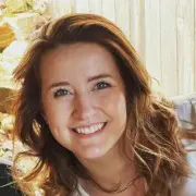 A person with wavy, shoulder-length brown hair and a bright smile is looking at the camera. The background features soft lighting and an abstract setting, possibly inside a Sunday salon, showcasing effortless style and charm.