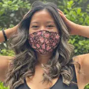 A person with long, wavy hair, wearing a black tank top and a patterned face mask, has their hands on their head. The background shows green foliage, evoking a refreshing escape one might seek after a visit to the local hair salon.
