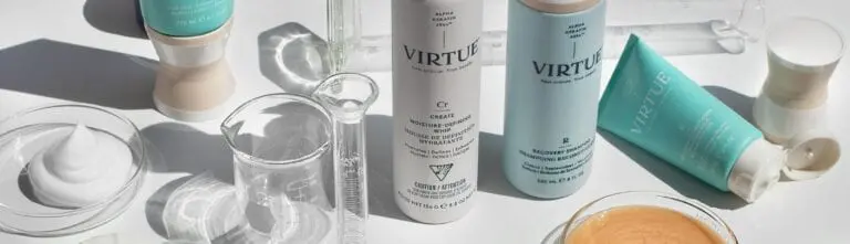 A collection of Virtue haircare products is displayed on a white surface, including bottles and tubes. Surrounding the products are clear laboratory glassware and a small dish containing a beige liquid. The scene has a clean, clinical aesthetic reminiscent of top-tier hair salons in Raleigh, NC.