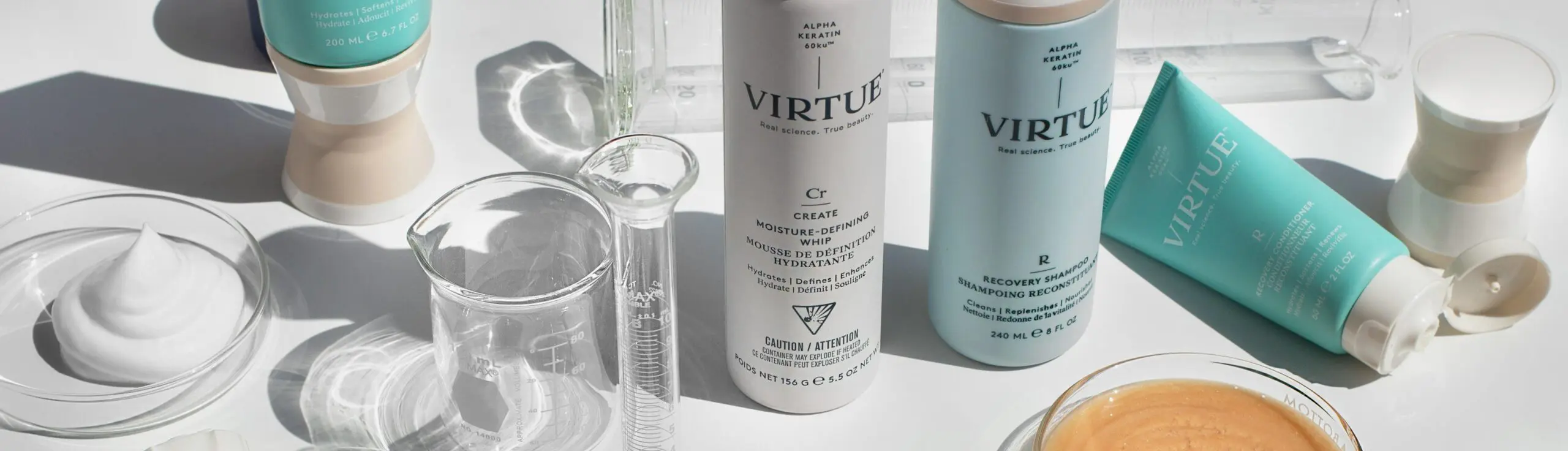 A collection of Virtue haircare products is displayed on a white surface, including bottles and tubes. Surrounding the products are clear laboratory glassware and a small dish containing a beige liquid. The scene has a clean, clinical aesthetic reminiscent of top-tier hair salons in Cary, NC.