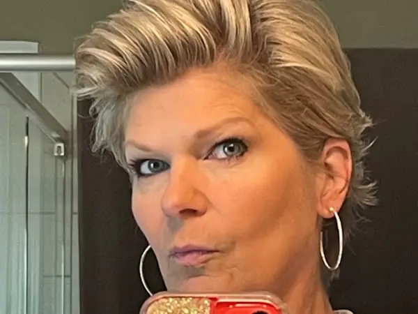 A person with short, blonde hair styled upwards, possibly groomed at the best hair salon in Cary NC, is taking a selfie. They are wearing large hoop earrings and looking into the camera with a neutral expression. The background features a glass shower and dark shower curtains. An orange phone case with glitter is partially visible.