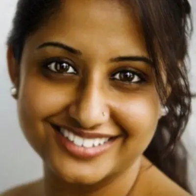 A close-up of a person with long dark hair tied back, smiling warmly at the camera. They are wearing pearl earrings and have a small nose ring. This could be the perfect look to get at your favorite hair salon.