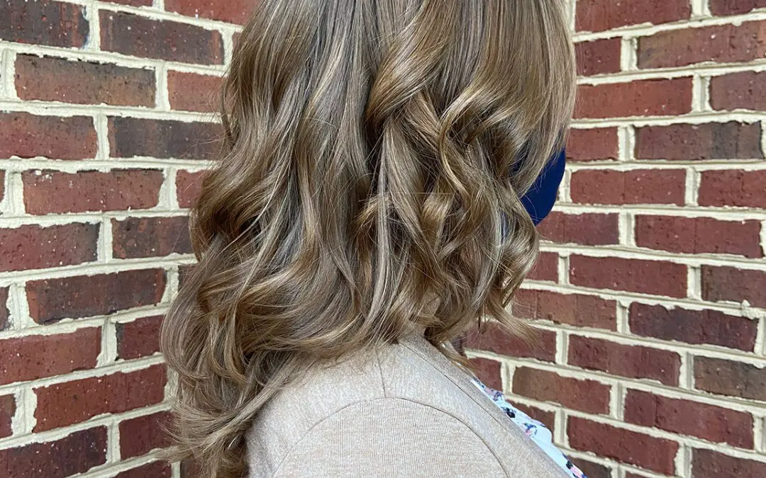 Side view of a woman with medium-length hair featuring dimensional caramel bronde highlights and voluminous, polished curls, created at Sunday Salon in Raleigh, NC.