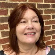 A woman with light skin and reddish-brown shoulder-length hair smiles slightly while looking to the side. She is standing in front of a brick wall, possibly having just left the best hair salon Cary NC offers.