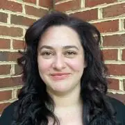 A person with long, wavy dark hair, freshly styled from a local hair salon near me, smiling at the camera. They are standing against a background of a brick wall and wearing a black top.