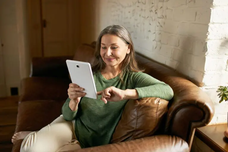 A woman with gray hair, wearing a green sweater, sits on a brown leather couch, smiling and looking at a tablet she is holding in her left hand. Her right arm rests on the couch armrest. The background features a light-colored brick wall and a doorway leading to the best hair salons in Raleigh, NC has to offer.