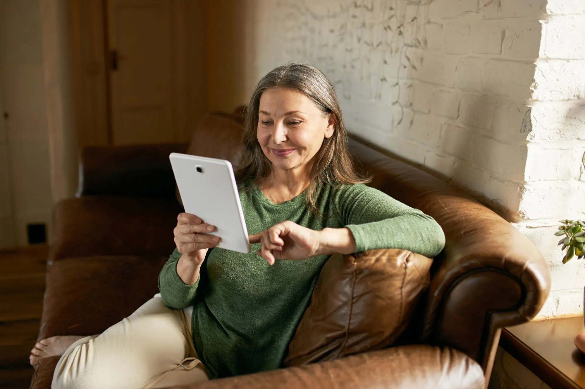 A woman with gray hair, wearing a green sweater, sits on a brown leather couch, smiling and looking at a tablet she is holding in her left hand. Her right arm rests on the couch armrest. The background features a light-colored brick wall and a doorway leading to the best hair salons Cary NC has to offer.