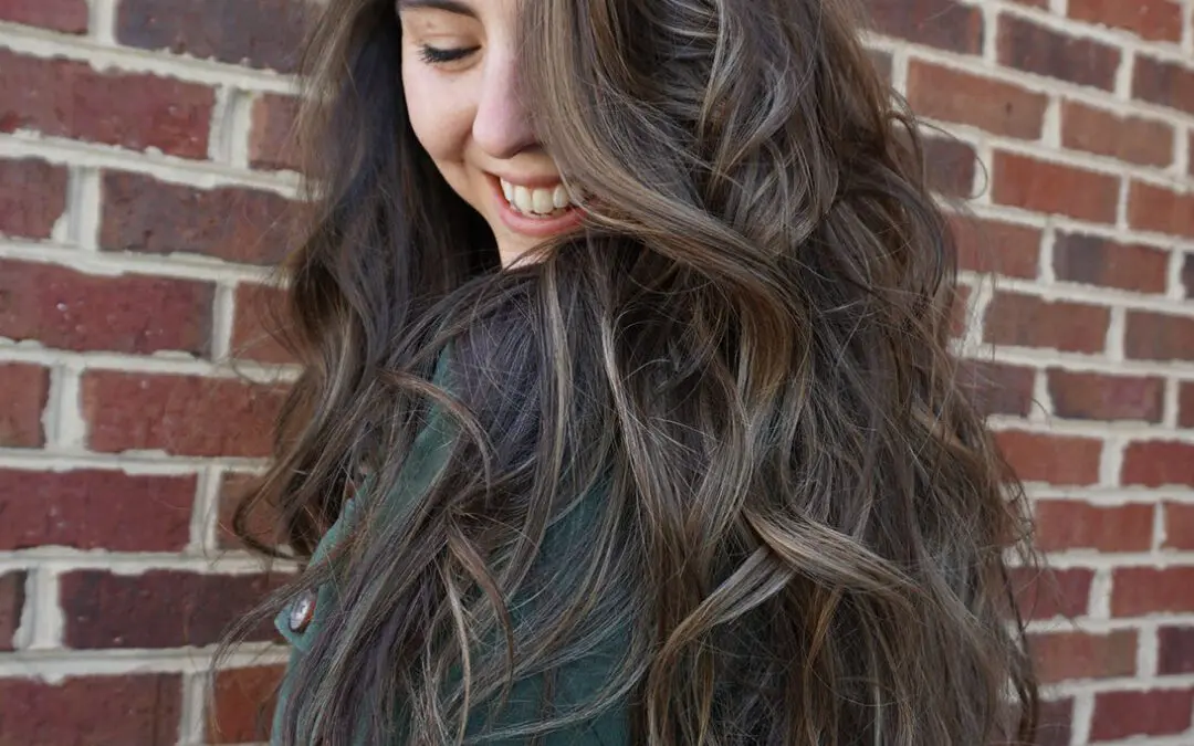 Woman smiling with ultra-long, thick brunette hair styled in voluminous cascading waves, featuring a subtle mushroom brown balayage, created at Sunday Salon in Raleigh, NC.