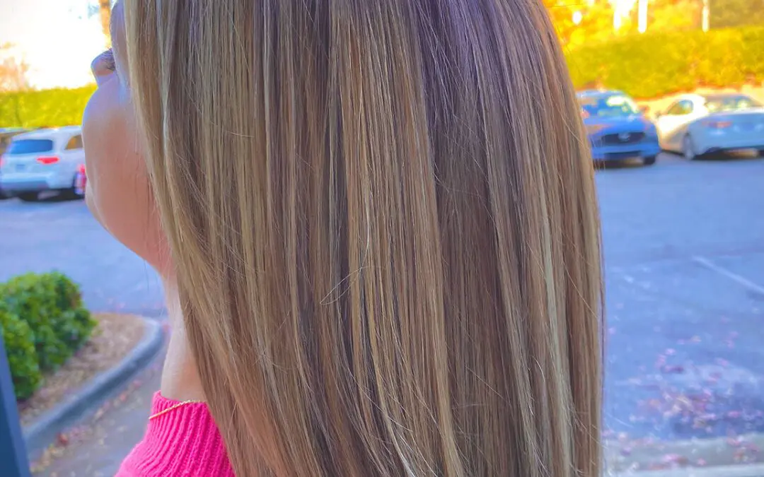 A person with long, straight, light brown hair stands outside, facing away from the camera. They are wearing a pink top. In the background, there are parked cars, trees with autumn foliage, and clear skies—perfect for a visit to the best hair salon in Cary, NC.