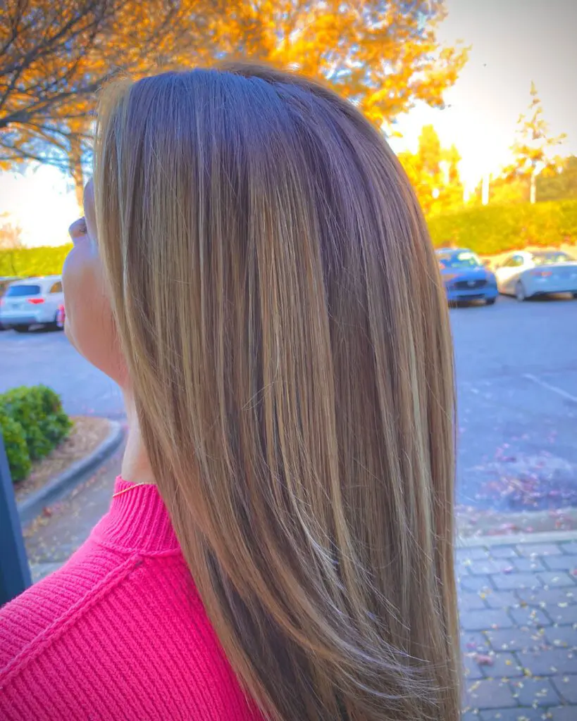A person with long, straight, light brown hair stands outside, facing away from the camera. They are wearing a pink top. In the background, there are parked cars, trees with autumn foliage, and clear skies—perfect for a visit to the best hair salon in Cary, NC.
