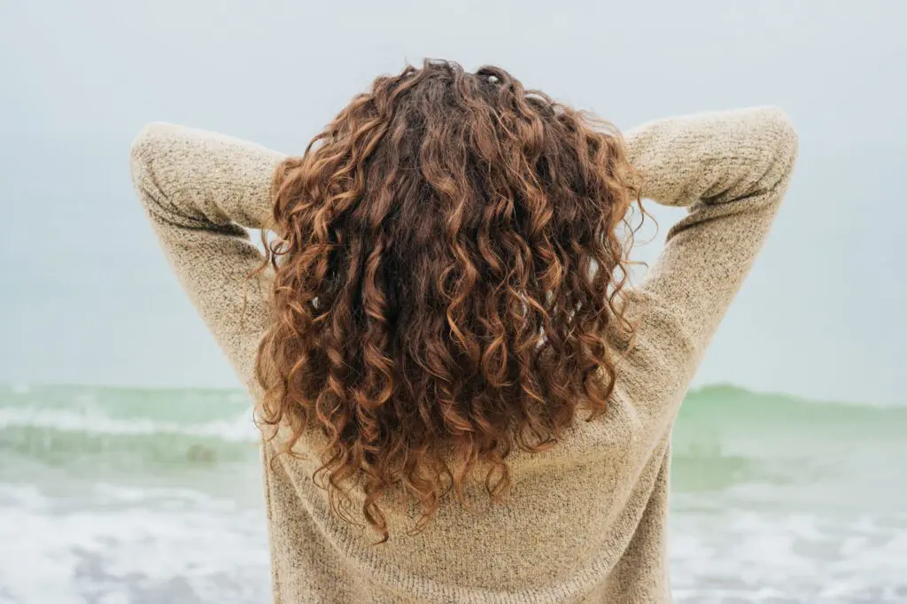 A person with curly, brown hair stands at the beach, facing the ocean with their arms raised and hands behind their neck. They are wearing a beige sweater. The sky is overcast, and the waves are gently rolling onto the shore, creating a serene backdrop for thoughts about visiting a hair salon soon.