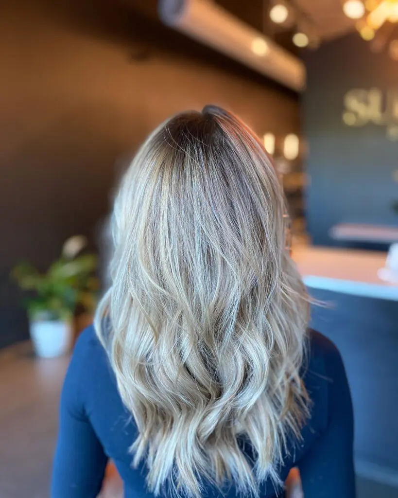 Back view of a woman inside a salon with long, wavy icy ash blonde hair featuring bright dimensional highlights and a dark shadow root by Lisa Edwards at Sunday Salon in Raleigh, NC.
