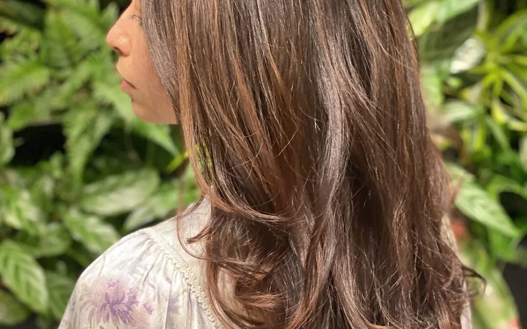 Side profile of a woman with long, thick hair featuring a deep chocolate brunette base and warm caramel highlights, styled in voluminous cascading layers at Sunday Salon in Raleigh, NC.