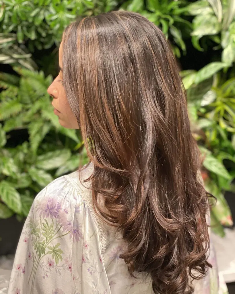 Side profile of a woman with long, thick hair featuring a deep chocolate brunette base and warm caramel highlights, styled in voluminous cascading layers at Sunday Salon in Raleigh, NC.