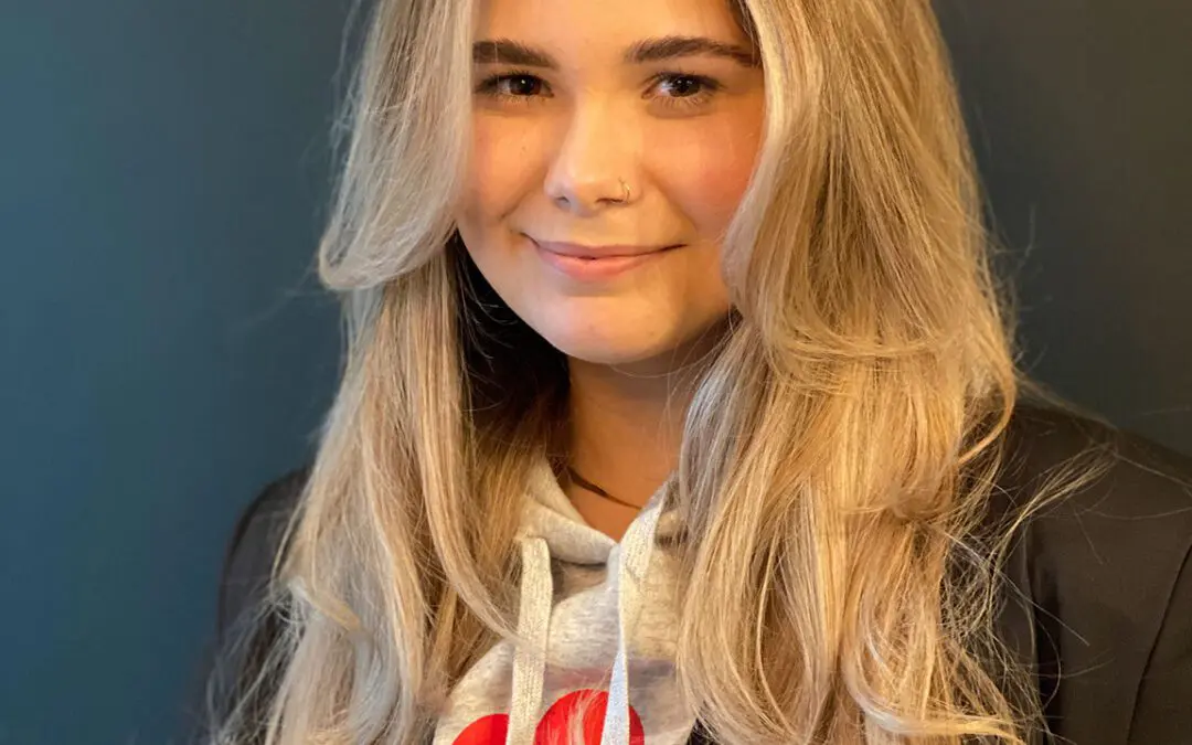 Smiling woman with ultra-long layered hair featuring a warm honey blonde balayage, bright face-framing highlights, and sweeping curtain bangs, styled at Sunday Salon in Raleigh, NC.