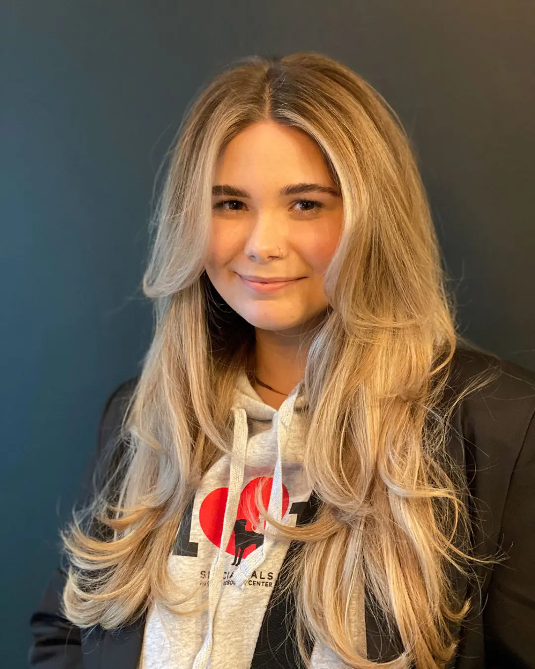 Smiling woman with ultra-long layered hair featuring a warm honey blonde balayage, bright face-framing highlights, and sweeping curtain bangs, styled at Sunday Salon in Raleigh, NC.
