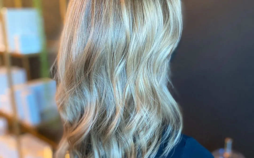 Back angle view of a woman with medium-long sandy blonde hair featuring dimensional highlights, textured waves, and a seamless shadow root at Sunday Salon in Raleigh, NC. Back view of a woman with shoulder-length bright lived-in blonde balayage, showing a pronounced shadow root and distinct textured waves by Sunday Salon Raleigh NC.