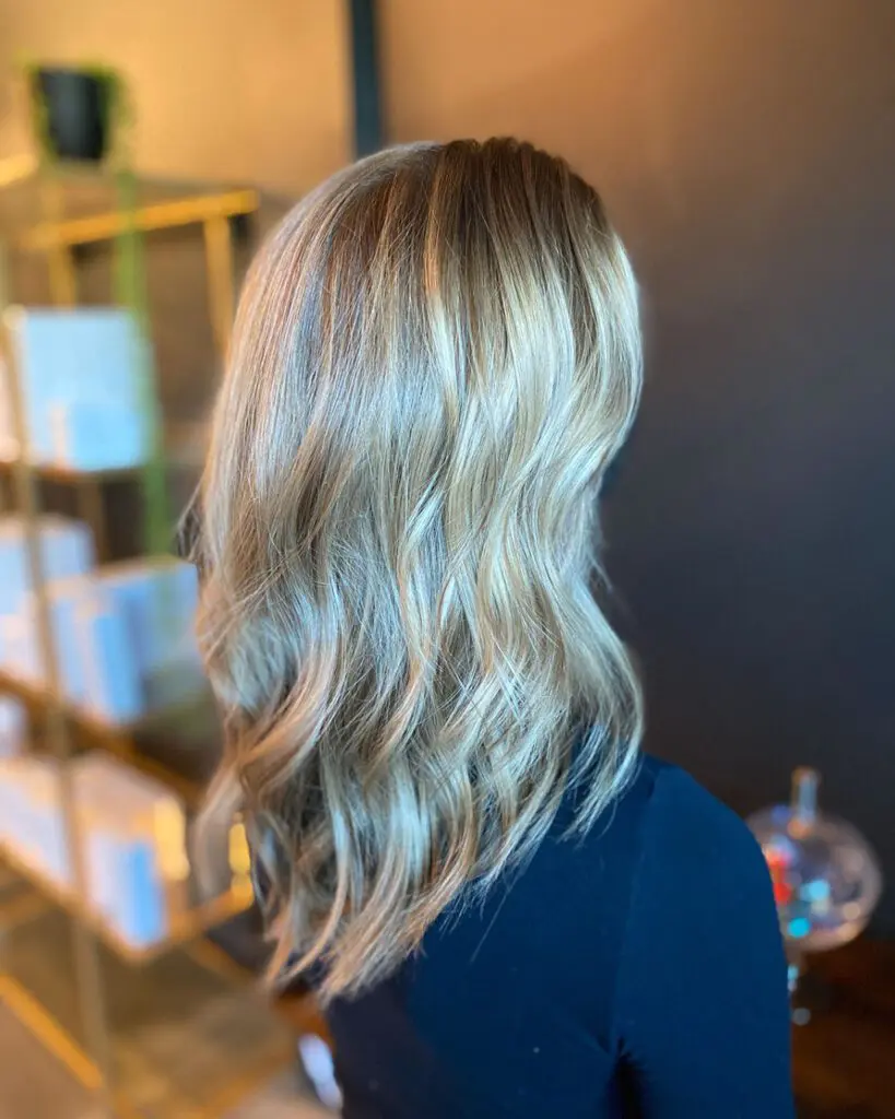 Back angle view of a woman with medium-long sandy blonde hair featuring dimensional highlights, textured waves, and a seamless shadow root at Sunday Salon in Raleigh, NC. Back view of a woman with shoulder-length bright lived-in blonde balayage, showing a pronounced shadow root and distinct textured waves by Sunday Salon Raleigh NC.