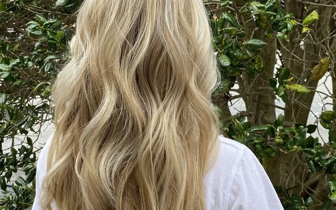 Back view of a woman outdoors with long wavy hair featuring bright creamy blonde highlights and a soft shadow root by Sunday Salon in Raleigh, NC.