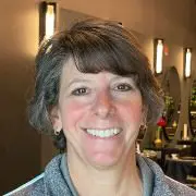 A person with short, grayish-brown hair is smiling at the camera. They are wearing a gray jacket and standing in a warmly lit indoor setting, perhaps one of the best hair salons Cary NC has to offer, with blurred background details including wall-mounted lights and some greenery.