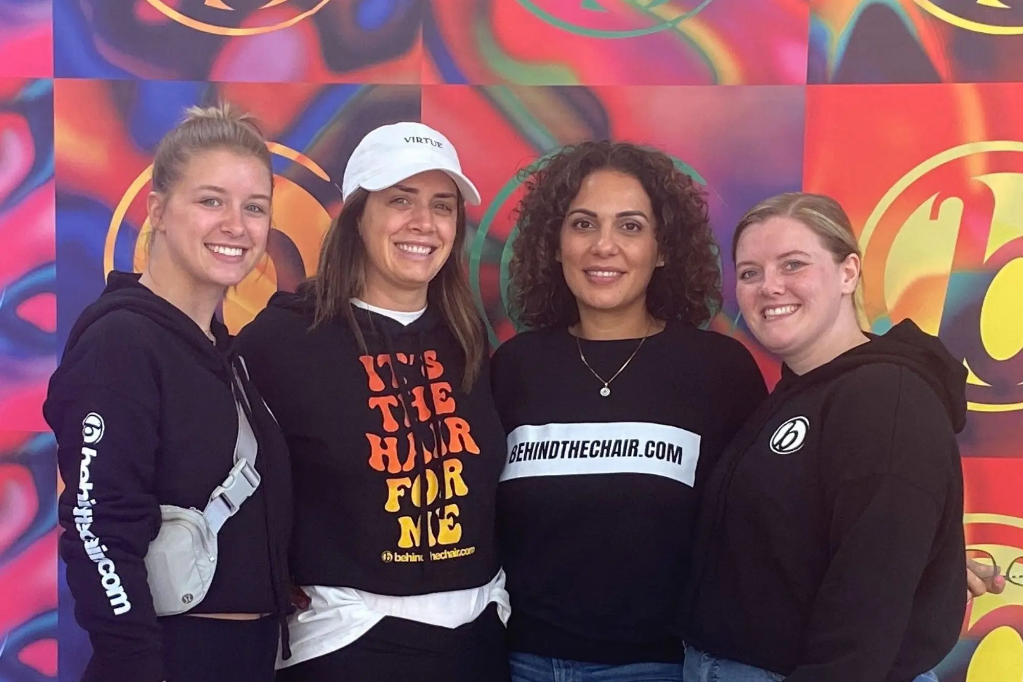 Four women are standing together and smiling against a colorful, patterned backdrop. They are dressed casually, with two wearing hoodies with the logo "BTC" and another in a hat. The central woman wears a shirt that reads "IT'S THE HAIR FOR ME," perfectly fitting for a Sunday salon outing at one of the best hair salons in Cary, NC.