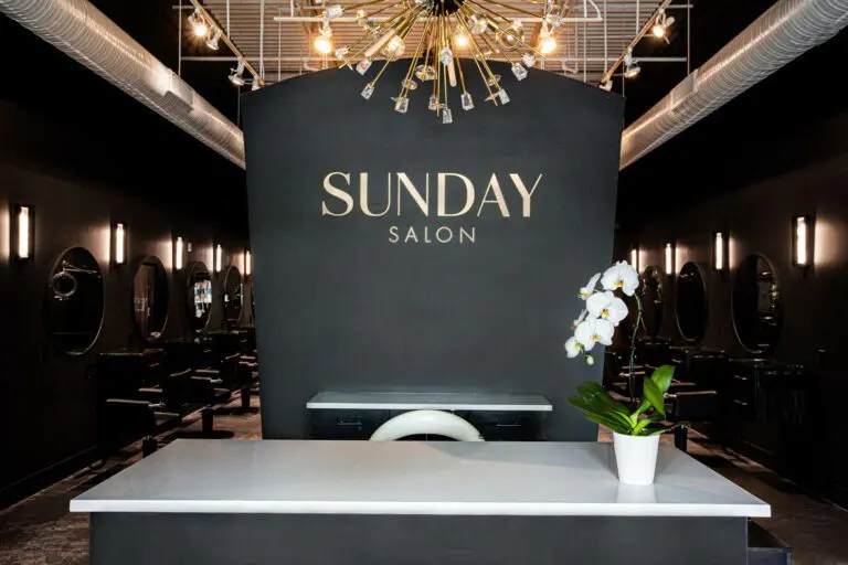 A modern, dimly-lit hair salon near me with a black color scheme, featuring the sign "SUNDAY SALON" on a black wall. A reception desk with a white surface and a potted orchid sits in the foreground. Circular mirrors and salon chairs are along the walls.