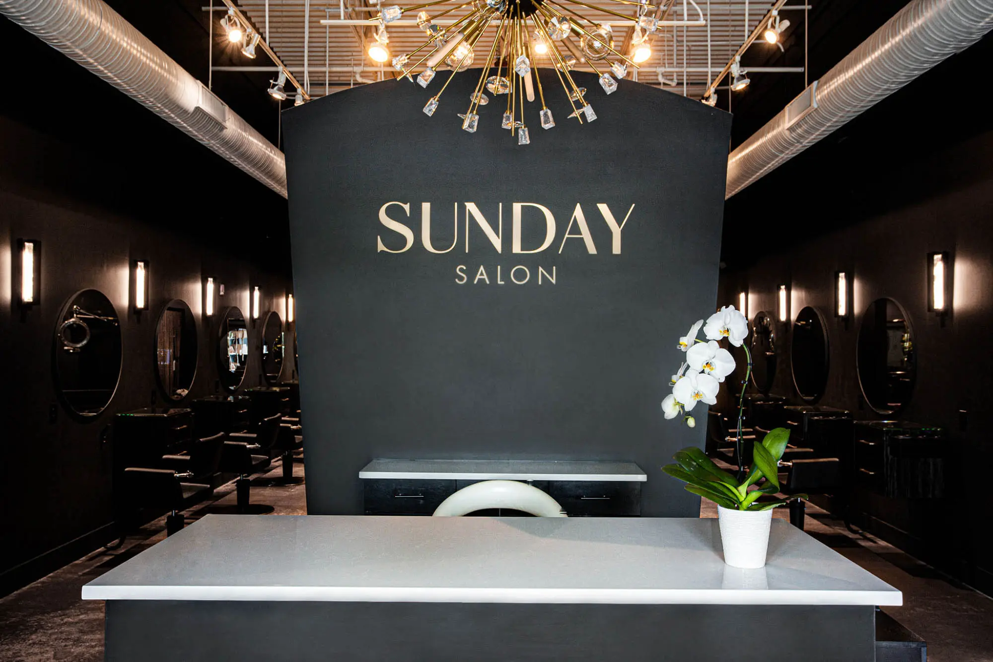 A modern, dimly-lit hair salon near me with a black color scheme, featuring the sign "SUNDAY SALON" on a black wall. A reception desk with a white surface and a potted orchid sits in the foreground. Circular mirrors and salon chairs are along the walls.