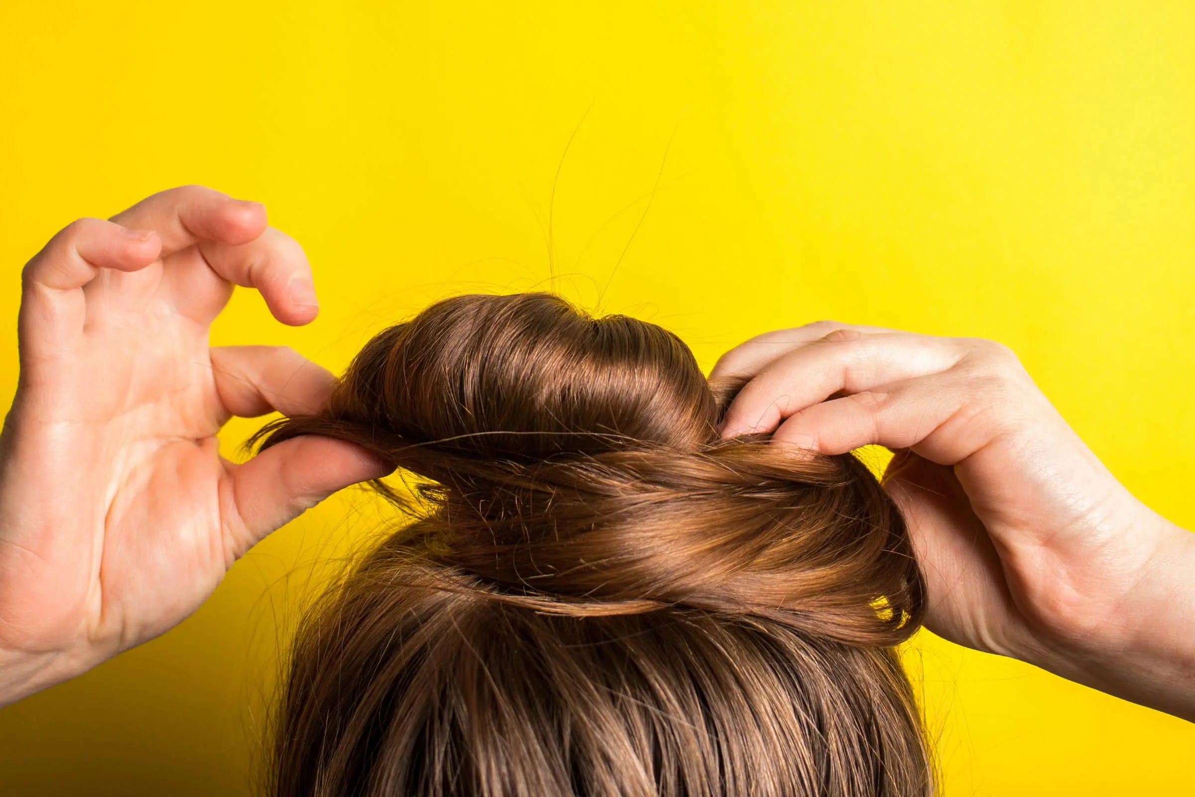 Close-up of hands arranging brown hair into a bun against a bright yellow background. The person is in the process of styling their hair, with fingers holding and shaping the bun, perhaps inspired by a visit to one of the best hair salons Cary, NC has to offer.