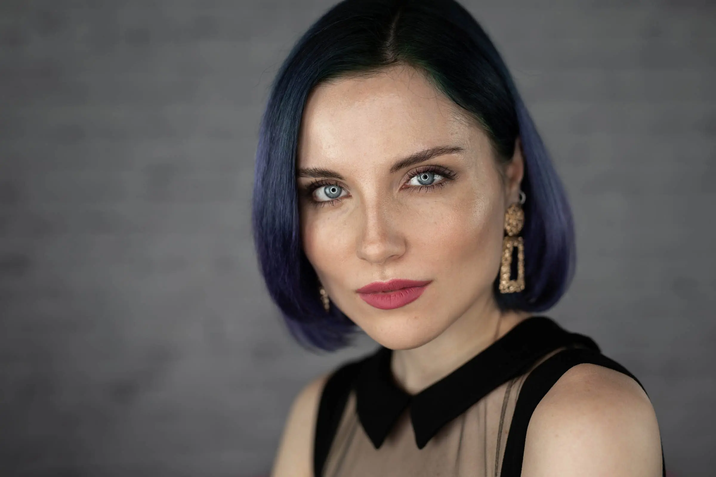 A person with short, straight, dark blue hair and fair skin is looking straight at the camera with a neutral expression. They are wearing a sheer black top with a solid black collar and large, ornate, rectangular earrings. Perfectly capturing the artistry you'd expect from the best hair salon in Cary, NC.