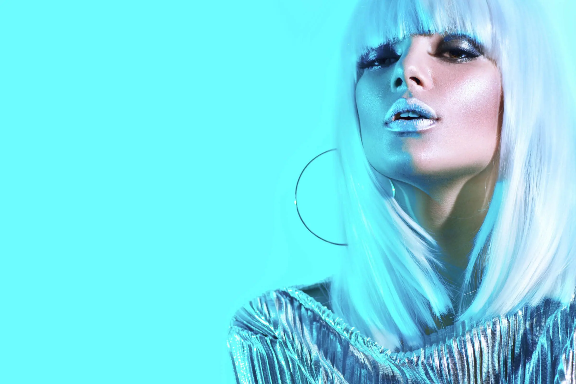 A person with platinum blonde hair and blunt bangs poses against a vibrant cyan background. They are wearing metallic silver clothing and large hoop earrings, with a confident expression and shimmering blue makeup, including blue lipstick. If you're seeking this bold look, visit a Sunday salon nearby.