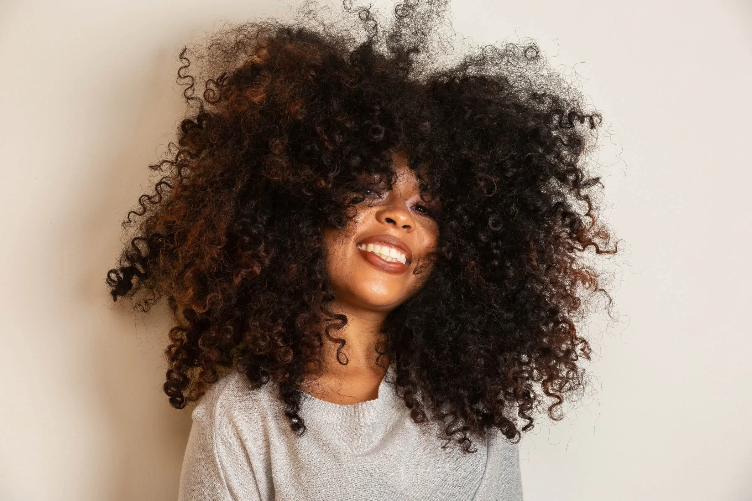 Smiling person with voluminous, curly hair that obscures part of their face, likely freshly styled at a hair salon. They are wearing a light grey top and are positioned against a plain white background.